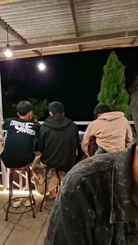 The video opens with a close-up of a young man from the shoulders up, seen from the side, wearing a dark, distressed denim jacket over a green t-shirt. He has short, dark hair styled with a fade on the sides and is looking off to his right. The background is blurred, but it appears to be an outdoor setting at night.

The camera then pans to the right, revealing a table with a phone, a green plate with what looks like skewers of food, and a glass with an orange beverage and ice. A white outlet is visible on a wooden railing in the background.

The camera continues to pan and zoom out, showing a wider view of the outdoor restaurant or cafe at night. Several groups of people are seated at tables. In the foreground, a group of young men are seated at a white table and benches. Further back, a two-story structure with an outdoor staircase lit by string lights is visible. More string lights are scattered throughout the outdoor seating area, illuminating the gravel ground and white fences. The overall atmosphere suggests a casual evening gathering at a dimly lit outdoor venue.