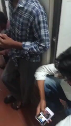 The video depicts a group of young men gathered in what appears to be a corridor. The lighting is dim, and the environment seems utilitarian, with tiled floors and walls. Several individuals are seated or squatting on the floor, some leaning against the wall. Many are holding and interacting with their mobile phones. One person in the foreground, wearing a plaid shirt, is looking down at their phone and holding it in their hand. Another individual, in a pink shirt, is also looking at their phone. The overall impression is one of a casual gathering, with the men engaged in activities on their phones, possibly socializing or passing time. The presence of discarded items on the floor suggests a lived-in space.