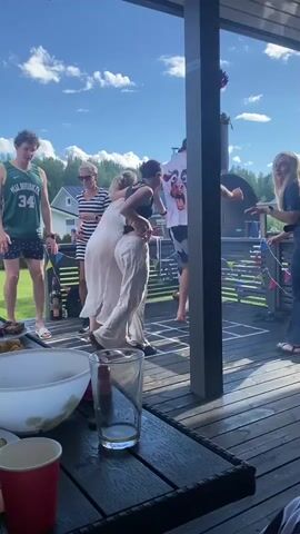 This is a vertical video clip showing a group of people dancing on a wooden deck outdoors. The scene appears to be a casual gathering, possibly a party or barbecue, on a sunny day. 

**Subjects:**
*   **People:** Several individuals are visible, both men and women, in various casual outfits. Some are actively dancing, while others are observing or in the background.
*   **Objects:** A table in the foreground holds a large white bowl, a red cup, a glass with liquid, and a bottle. In the background, a barbecue grill is visible, along with some colorful bunting strung across a railing. There's also a basketball jersey with the name "MILWAUKEE" and the number "34" on the back of one person.
*   **Scene/Setting:** The setting is an outdoor wooden deck with a railing. Beyond the deck, there's a lawn with trees and houses, suggesting a residential area. The sky is bright blue with fluffy white clouds.

**Activity/Event:**
The primary activity is dancing. The people are engaged in energetic movement, suggesting music is playing and they are enjoying themselves.

**Notable Details:**
*   **Time of Day:** The bright sunlight and clear blue sky indicate it is daytime, likely midday or afternoon.
*   **Weather:** The weather appears to be warm and pleasant, suitable for an outdoor gathering.
*   **Location Cues:** The presence of a barbecue, casual attire, and the general ambiance suggest a summer celebration. The prompt specifies Joensuu, Finland, which aligns with a potential summer event.
*   **Emotions:** The overall mood seems joyful and relaxed, with participants appearing to have fun.

**Visible Text:**
The most prominent text visible is "MILWAUKEE" and the number "34" on the back of a green basketball jersey worn by one of the men.