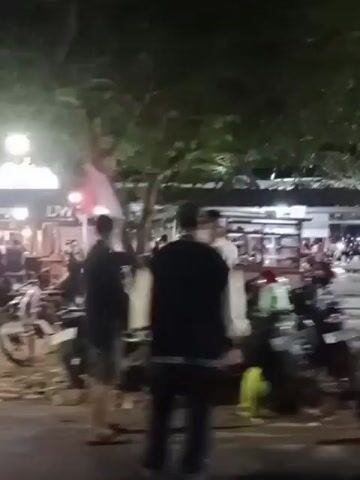 The video shows an outdoor gathering at night, likely a street food market or a casual dining area. Several food stalls are visible, illuminated by string lights and lamps. People are seated at tables, some eating and socializing. Motorcycles are parked nearby, suggesting a mode of transportation for some patrons. A flag, possibly red and white, is hanging from a pole. The overall atmosphere appears relaxed and lively.