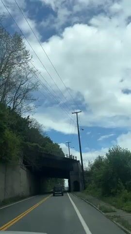 The video shows a car driving on a two-lane road passing under a concrete bridge. The sky is partially cloudy with overhead power lines and telephone poles next to the road. Trees border both sides of the road.
