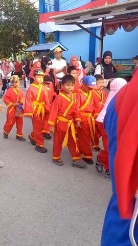 The video shows a group of young children, dressed in red and yellow martial arts uniforms, walking in a procession. They are accompanied by adults, some of whom are also wearing headscarves. The children appear to be participating in an event, possibly a parade or a school activity. The environment seems to be outdoors, with a building visible in the background, decorated with flags and banners. The overall mood appears to be festive and organized.