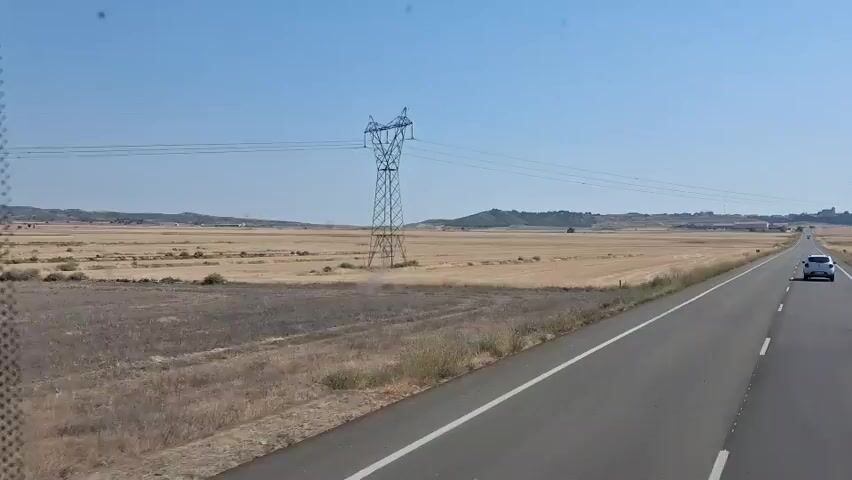 This is a wide shot of a rural highway on a clear, sunny day. The asphalt road stretches into the distance, with a white car driving away from the camera in the right lane. On the left side of the road, there is a vast, dry, golden field. In the distance, a large, green electrical tower stands prominently, with power lines stretching across the horizon. The sky is a clear, vibrant blue. The landscape appears to be arid and agricultural. The camera is in motion, likely from a vehicle also on the road, following the white car.