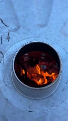 The video shows a close-up of flames burning inside a metal cylindrical opening on a large, light-colored metal surface. The flames are orange and yellow, and some smoke is visible. The metal surface is slightly textured, and some dark, indistinct markings are present.