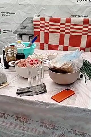 A table is set with various items, including bowls of food, bottles, and a blender. In the background, a banner with images of houses and text is displayed. The scene appears to be outdoors, with wooden flooring visible beneath the table. A person's hand reaches into a bowl of food on the table, suggesting preparation or serving. A smartphone is also present on the table, along with a checkered cloth.
