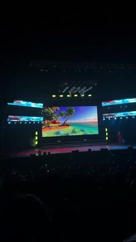 The video shows a dark auditorium filled with an audience facing a stage. A large screen on the stage displays a tropical island scene with a palm tree and clear blue water. There are other smaller screens on either side.  A woman in a pink outfit stands on the stage. Overhead, there are lighting rigs and a series of white balloons. Blue lights are shining down onto the stage from above.