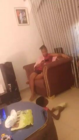 A woman sits on a brown armchair in a room. She holds a smartphone in her hands and appears to be looking at it.  A child lies on the floor near a round, blue table. The table has various items on it, including a package of diapers.  A speaker is visible against the wall.  There's a curtain behind the woman and a framed picture on the wall.  The room is well-lit.