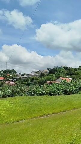 The video pans across a rural landscape under a cloudy blue sky. In the foreground, a lush green field is visible, with a subtle slope. Beyond this, the land rises into a series of hills dotted with trees and vegetation. Various buildings, including houses and what appear to be greenhouses or agricultural structures, are scattered across the hillsides. The overall impression is one of a peaceful, verdant, and sparsely populated rural area.