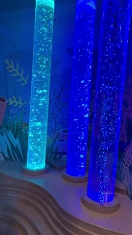 Three tall, clear cylindrical tubes filled with water and illuminated by colored lights stand vertically on a wooden base. Bubbles rise from the bottom of each tube, creating a dynamic visual effect. The tubes are lit with blue light, with the left tube transitioning to yellow at the bottom. The background features a blue wall with a mural depicting underwater plant life in shades of pink, purple, and white. The floor is a light wood material with a wavy, layered design. The overall lighting is dim, with the illuminated tubes being the primary light sources. The camera is positioned at a medium angle, looking slightly up at the tubes. The scene appears to be in a dimly lit indoor space, possibly a sensory room or a children's play area.
