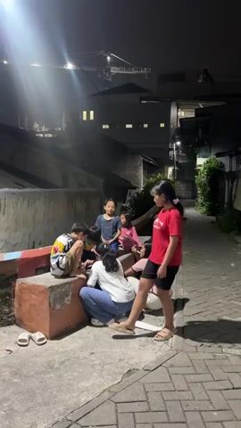 A group of children are gathered on a concrete area at night. The children are sitting and standing around a low, orange-colored raised platform. One child in a red shirt is standing and looking down at the other children. Two children in white and blue clothing are seated on the ground. One child in a white shirt is facing the viewer and looking down. Another child in blue pants is facing away from the viewer and looking down. Three other children are seated on the orange platform, one wearing a colorful shirt, and two wearing pink shirts. They are all looking at something on the ground. The background shows buildings and a street with a brick-like pattern. The lighting is dim, with a bright light source to the left of the frame. A crane is visible in the distance.