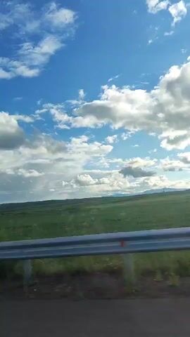 The video captures a scenic view of a green field under a vast blue sky filled with white, puffy clouds. A metallic guardrail runs horizontally across the foreground, separating the viewer from the field. The camera appears to be in motion, possibly from a vehicle, as the landscape unfolds. The overall impression is one of a bright, clear day with a sense of open space.