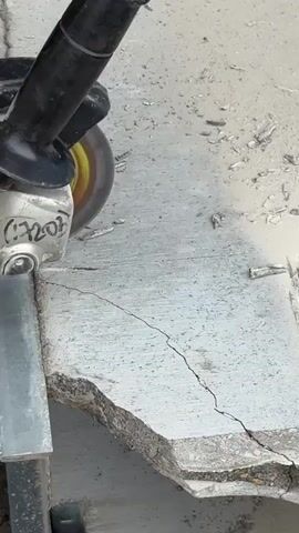 The video shows a close-up of an angle grinder with a rotating cutting disc being used to cut a concrete slab. The concrete slab is cracked and broken. The angle grinder is held by a black handle and has a blue casing with some writing on it, including what appears to be "C7207". The cutting disc is creating sparks as it grinds away at the concrete. Small pieces of concrete are flying off as the cut progresses. The background is blurred, but appears to be a light-colored surface, possibly more concrete or pavement. The lighting suggests it is daytime. The video is very short, showing only the action of cutting.