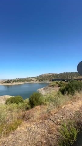 A landscape shot of a large body of water, possibly a reservoir, on a clear, sunny day. The water is calm and reflects the bright blue sky. The surrounding terrain is dry and covered in sparse vegetation, including grasses and shrubs. In the distance, there are hills and what appears to be a dam or bridge structure. The camera pans slowly to the right, revealing more of the shoreline and the expansive sky. The overall impression is one of a natural, somewhat arid environment bathed in bright sunlight.
