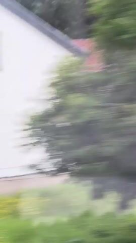 The video shows a panning view of a street in a residential area. A white house with a green roof is visible on the left side of the frame. A red-roofed building is in the foreground. A black SUV is driving past a row of garages on the right side of the frame. The environment appears to be a suburban or rural setting, with trees and vegetation present. The overall impression is one of a normal day in a quiet neighborhood.