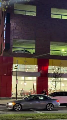 The video shows a street scene at dusk or night. A multistory brick building with yellow and red accents dominates the background, with lit windows on several levels. A parking structure appears to be integrated into the building, with cars visible in the upper levels. A restaurant with a bright red and yellow sign, possibly "In-N-Out Burger," is visible on the ground floor. People are seen inside the restaurant and walking on the sidewalk. Cars are driving on the street in the foreground, and there are trees lining the sidewalk. The overall impression is a busy urban environment.