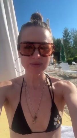 The video begins with a close-up selfie of a woman with her hair in a bun, wearing large, tortoiseshell sunglasses and a black bikini top with a daisy design. She is on a beach, with sand and lounge chairs visible in the background. She smiles at the camera. The camera then pans slightly to the right, revealing another woman sitting next to her, also wearing a bikini and sunglasses. The first woman then turns back to the camera and makes a kissing face. She speaks briefly, and then the camera pans again to show both women, with the second woman smiling. The video continues with shots of the beach scene, including lounge chairs, umbrellas, and other people in the background. The first woman is visible in some shots, either alone or with the second woman. The overall impression is of a casual day at the beach.