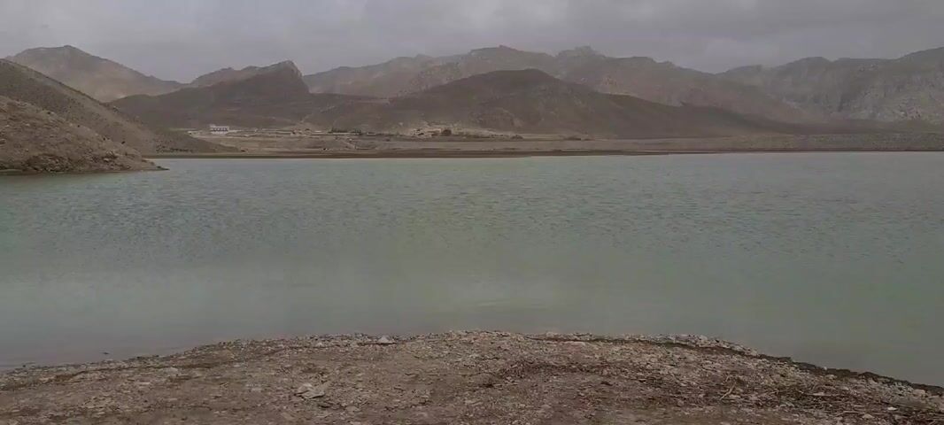The video shows a lake surrounded by mountains under a cloudy sky. The water is a light greenish-gray color with gentle ripples on the surface. The shoreline is made up of rocky, brown soil. In the background, there are several mountains with a barren, rocky appearance. The sky is overcast, and the overall scene appears calm and still. A small structure is visible on the shore in the distance. There is some visible movement in the water toward the end of the video.