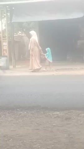 A woman wearing a long beige dress and a cream headscarf walks hand-in-hand with a small child wearing a blue headscarf. They are walking on a paved road in front of a building with an archway. The archway has text on it that is difficult to read due to the image quality. There is a trash can next to a sign with "RW 02" on it near the archway. The environment appears to be dusty or hazy.