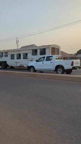The video shows a street scene. The initial shot features a white Toyota pickup truck parked next to a dilapidated, white trailer. A man in an orange shirt is walking toward the trailer. The camera then shifts to the street, where a black Kia and a silver car pass by. Following this, a yellow tricycle, also known as a "Keke," comes into view, approaching the camera. The video ends with a view of a young man in the tricycle looking directly at the camera, while a few people stand nearby watching other vehicles. The environment appears to be a road in a city or town. The lighting indicates it is late afternoon or early evening. There are various vehicles parked along the side of the road, including a large truck, a Mercedes, and the Toyota pickup truck seen earlier.  There are telephone poles and power lines present in the background.