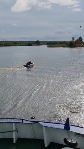 The video shows a body of water with a small inflatable boat with two people in it moving away from the camera. The camera is positioned on a larger boat, with its railing visible in the foreground. The water is choppy, with a visible wake behind the inflatable boat. In the background, there is a tree-lined shore with some structures. The sky is overcast with some patches of blue. There are birds flying in the sky. The time stamp indicates the video is very short, only a few seconds.