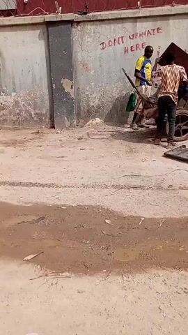 The scene shows two men tending to a wheelbarrow near a wall. The wall is painted with peeling paint and has the graffiti message "DON'T URINATE HERE IN." One man, wearing a yellow and blue shirt, stands by the wheelbarrow, holding a long stick. The other man, clad in a patterned shirt and dark jeans, bends over the wheelbarrow, appearing to adjust its contents. The wheelbarrow is filled with assorted debris, including what looks like discarded leaves. In front of the wheelbarrow, several dark, flat objects are scattered on the ground. The ground around them is a mix of dirt and gravel, with some damp patches.