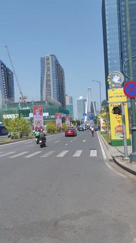 The video shows a daytime urban scene in what appears to be a city in Southeast Asia. The camera is positioned at street level, looking down a multi-lane road. Several modern high-rise buildings dominate the skyline. Construction is evident, with cranes and scaffolding surrounding some buildings. The road has white directional arrows painted on the pavement. Traffic consists mainly of motorbikes and a few cars moving in both directions. Banners with Vietnamese text and images are displayed along the roadside. The sky is clear and blue. The scene gives the impression of a bustling, developing urban environment.