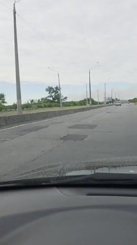 The video shows a first-person view from inside a vehicle driving on a paved road. The road has multiple lanes and is lined with streetlights and a concrete barrier. Beyond the barrier, there is vegetation and some buildings in the distance. The sky is overcast. Several other vehicles are visible on the road ahead. There are some visible cracks and imperfections on the road surface. In the distance, a billboard is visible.