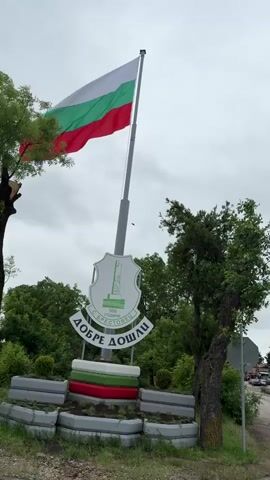 The video shows a view of a Bulgarian flag waving in the wind, mounted on a tall, silver pole. Beneath the flag, a sign welcomes visitors to Brestovets. The sign is white with green detailing and features an image of a monument. The words "Добре Дошли" (Welcome) are prominently displayed in a curved design. Underneath the sign is a small landscape bed constructed from concrete blocks, painted with the colors of the Bulgarian flag: white, green, and red. The scene is surrounded by trees with green foliage, and the sky is overcast with gray clouds. A street and several parked cars are visible in the background.