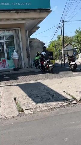 The video shows a street scene. In the foreground, a gray asphalt road with a concrete gutter is visible, casting a shadow from an unseen object. A paved sidewalk runs parallel to the road, with a concrete drain cover. On the sidewalk, a person wearing a helmet and a green vest is standing next to a motorcycle, interacting with another person. The motorcycle is black and white with a rider wearing a helmet and a white shirt. Behind them, a building with a glass storefront displays a large ice cream cone statue. A sign above the building reads "CREAM, TEA & COFFEE". To the right, a yellow kiosk or stand is present, with more motorcycles parked nearby. Trees and buildings are visible in the background under a clear blue sky. Power lines crisscross the scene. The overall impression is of a commercial area on a sunny day.
