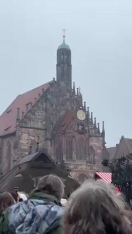 The camera follows a crowd of people walking toward two monumental structures: a tall church with a large clock and a decorative tower with intricate sculptures. The people in the crowd are wearing winter clothing and walking in a line. One person is taking a photo of the tall decorative tower. The scene appears to be outdoors on an overcast day. Some small buildings with red-and-white striped roofs can be seen between the people and the tower.