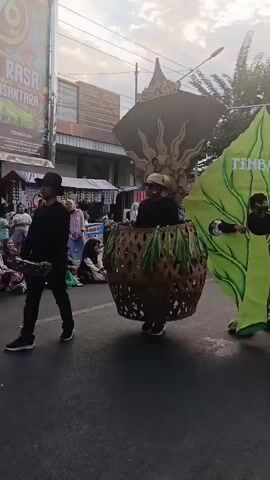 The video shows a street parade with several people in costumes. In the foreground, a person in a large basket-like costume decorated with green leaves walks down the street. To the right, another person is wearing a giant green leaf costume with the word "TEMBAKAU" written on it. This person is giving a thumbs up. Behind them, other participants and spectators are visible, along with buildings and streetlights. The atmosphere appears festive.