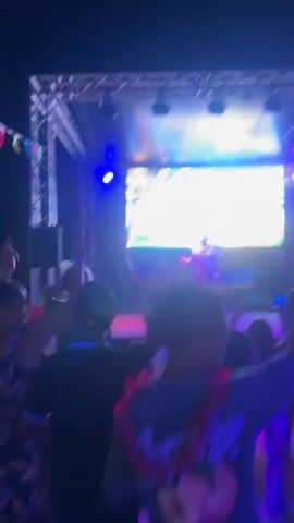 The video shows a crowd of people dancing in front of a stage at night. The stage has a large screen displaying abstract visuals, and a DJ is visible on the stage. Blue and green lights illuminate the scene, and some people in the crowd are holding glowing sticks. The overall atmosphere appears to be a lively outdoor party or concert.