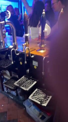 The video is taken from behind a wooden bar in a pub, focusing on the beer taps. The taps are silver with wooden handles. Behind the bar, a group of people is dancing with their arms raised. The lighting changes colors, cycling through green, blue, and purple. The pub has a rustic, old-fashioned feel, with various decorations hanging from the ceiling, including what appear to be old mugs or tankards. Pictures and possibly some signs are hanging on the walls. The overall atmosphere seems lively and festive.