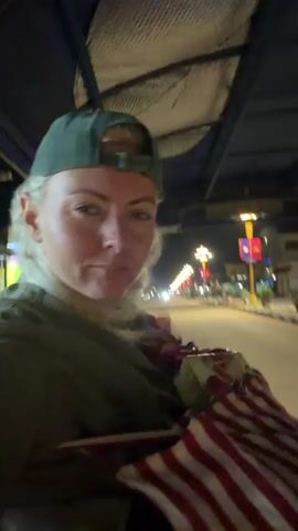 This is a close-up, first-person perspective video taken at night from inside a moving vehicle, likely a tuk-tuk. In the foreground, a woman wearing a green baseball cap and a green hoodie is visible. She is looking directly at the camera. To her right, in the middle ground, a man wearing a light-colored shirt, a scarf, and a light-colored baseball cap is visible. He is looking towards the camera and holding up two fingers in a peace sign. The background shows a street scene with buildings, streetlights, and other vehicles, indicating an urban environment. The vehicle interior is dark, with a roof visible overhead. The woman is holding a red and white striped bag or blanket. The man is also holding a bag or blanket.