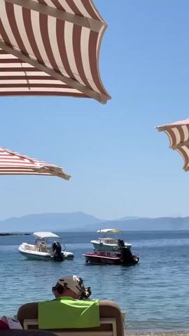 A serene beach scene unfolds under a bright blue sky. Striped umbrellas, shades of red and white, cast dappled shadows on the sandy shore. Three small boats are anchored in the calm, clear blue water, with two in the foreground and one further back. In the middle ground, a man in red shorts walks along the beach. In the background, hazy mountains rise against the horizon. The foreground features a beach chair with a person in it, facing the water, and a person in a swimsuit with arm floaties.