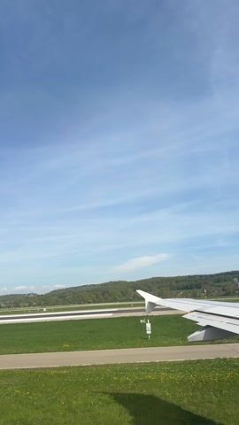 The video shows a view from inside an airplane, likely near a window. The aircraft is on a runway surrounded by green grass and dandelions. In the distance, a forested hill stretches across the horizon under a bright blue sky with scattered clouds. A portion of the plane's wing is visible in the lower right corner of the frame.