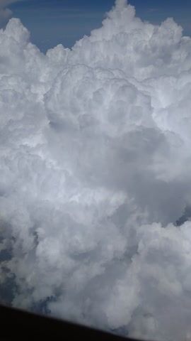 This media file captures a view from an airplane window, showcasing a vast expanse of clouds against a deep blue sky. The clouds are predominantly white and fluffy, with some areas appearing darker and more shadowed, suggesting varying altitudes and densities. The scene is dominated by these atmospheric formations, creating a sense of depth and grandeur. There are no visible people or distinct objects within the frame, the focus being entirely on the natural phenomenon of the clouds.

The time of day is difficult to determine precisely, but the bright blue sky and the illuminated clouds suggest it is daytime, likely mid-morning or afternoon, as there are no signs of dawn or dusk. The weather appears to be partly cloudy, with the potential for cumulus or cumulonimbus clouds given their size and texture. The angle of the shot, looking downwards and outwards, is typical of an aerial perspective from a commercial flight.

The overall mood conveyed is one of serenity and awe, the overwhelming presence of the clouds evoking a feeling of being high above the world. The image is oriented such that the sky is on the left and the clouds fill the majority of the frame, with a dark, rounded edge on the right suggesting the airplane's window frame. There is no visible text in the media file. The location context of Ilidža, Bosnia and Herzegovina, implies that this view is from a flight traveling over or near this region.
