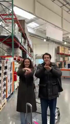 A young man and woman are dancing in the aisle of what appears to be a Costco store. The woman is wearing a long, dark green coat and has curly brown hair. She also has a purple backpack on. The man is wearing a black puffer jacket and jeans. They both have smiles on their faces and are dancing together in sync. There is a shopping cart in front of them. The store has tall shelves stocked with products. The floors are concrete. Overhead lights illuminate the space.