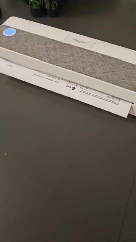 A white shredder sits on a dark grey surface. A sheet of paper with text and a barcode is being fed into the shredder from the right side. The paper is partially inside the shredder, with the text and barcode visible. A blue light is illuminated on the top of the shredder, indicating it is in operation.
