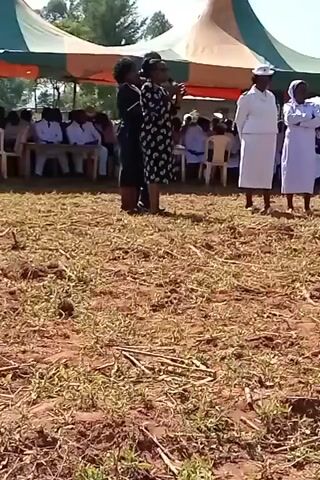 This video shows a group of people gathered outdoors. Underneath a large tent, people are seated in rows of chairs. In front of the seated group, three women stand in a line. The woman in the middle is speaking into a microphone. She is wearing a patterned dress. The woman to her left wears a dark dress. The women to the right are wearing white uniforms, including hats. The ground is dry, with patches of green grass.