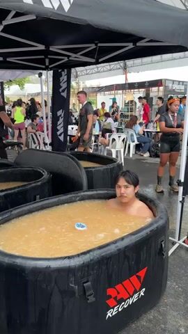 The video shows a man sitting in an inflatable ice bath tub. The tub, marked with the word “RECOVER” and another undecipherable word, is filled with water and ice cubes, with a thermometer floating on the surface. He is shirtless with short dark hair. Other people are nearby, some sitting at tables and chairs, others standing. Behind them is a booth with the words “SPARTAN THAILAND” visible. There are black flags with white writing. A man walks by the side of the tubs carrying a cup. The video appears to be taken at an outdoor event.