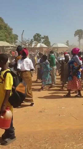 The video shows a crowd of people, mostly women and children, gathered in a dusty, rural environment. Many of the women are wearing brightly colored, traditional clothing, including headwraps and patterned skirts or dresses. Some are carrying babies or young children on their backs. Several individuals are wearing white shirts with visible writing on them. The crowd is spread across a sandy or dirt-covered area, with simple buildings and trees visible in the background. There's a general sense of movement and activity, suggesting some sort of gathering or event. Dust kicks up in the air as the crowd moves. The sky is blue and clear.