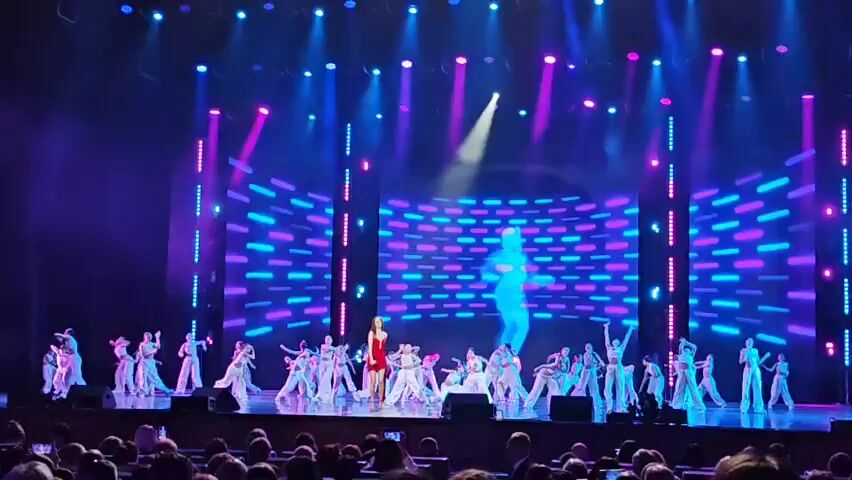 A group of dancers in white costumes performs on a stage with a large screen in the background displaying a pixelated image. A singer in a red dress stands center stage, singing into a microphone, while a silhouette of a dancer is projected onto the screen behind her. The stage is lit with blue and pink lights, and spotlights illuminate the performers. The audience is visible in the foreground, seated in a theater.
