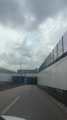 A driver is approaching a dark tunnel entrance on a highway. The weather is overcast, with a sky filled with dark grey clouds. There is a white concrete wall with a green fence running along its top on the right side of the road. A grey concrete wall with a fence runs along the left.  Above the tunnel entrance is a sign indicating a speed limit of 60 km/h. Three large ventilation fans are visible inside the tunnel entrance. Green lights are on, indicating that the tunnel is open.