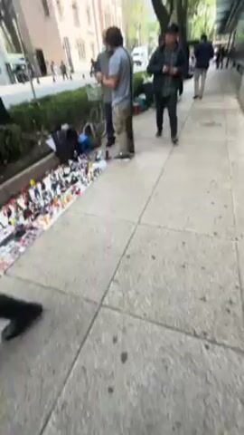 The video shows a sidewalk scene in what appears to be a city. A man in a grey t-shirt and khaki cargo pants is looking at a phone. Next to him, another man in a blue shirt is also looking at a phone. They are standing near a collection of toys laid out on a white sheet on the ground, likely for sale. A man in a flannel jacket is walking away from the group, and other people are walking by in the background. A bicycle is parked nearby, and there are buildings and trees in the background.