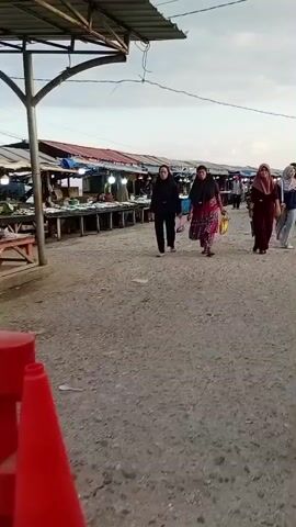 The video shows a group of women walking down a gravelly, open-air market. The women are dressed in hijabs and traditional clothing. One woman in the foreground is wearing a maroon dress and a light pink hijab, carrying a dark bag. To her left, another woman is wearing a black hijab and a patterned dress, carrying a yellow bag. Further to the left, a third woman in a black hijab and black clothing walks with a pink bag. The market stalls are visible on either side of the path, some displaying goods. The sky is overcast, suggesting it is either early morning or late afternoon. The ground is covered in small stones and dirt. The overall impression is a casual stroll through a bustling marketplace.