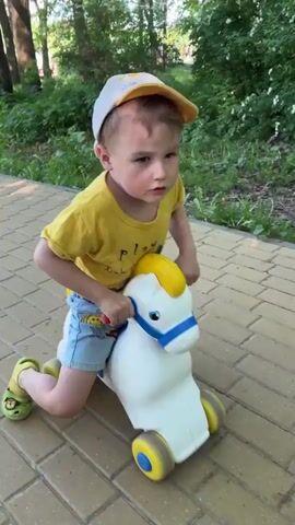 A young child wearing a yellow t-shirt, blue shorts, a yellow and white cap, and green clogs is riding a white plastic toy horse with yellow and blue accents on a brick paved walkway. The child is pushing off the ground with their feet to propel the horse forward. Green foliage is visible in the background.