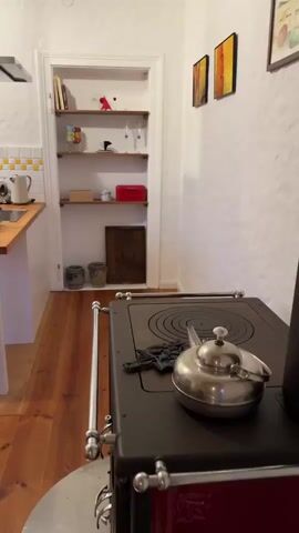 The video pans across a rustic kitchen. To the left, a stainless steel range hood hangs over a stovetop with a kettle on it. Adjacent is a wooden countertop with a blue pitcher and a yellow plate. Further left, a red cabinet is visible. The kitchen features white tiled backsplash and white textured walls, adorned with two framed artworks. White open shelves display various kitchen items. A white island with a trash can is situated in the foreground. To the right, a tall wooden cabinet with glass doors stands against a wall. Beyond it, a large window reveals a garden and a blue sky. The floor is made of wooden planks. A radiator is visible beneath the window. Two trash cans are placed near the radiator. A coffee maker sits on a small wooden cart near the window.