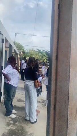 The video begins with a close-up of a metal pole. It then pans to a group of young people, mostly female, standing outside a building. Some are wearing white shirts with writing on them, and one person is holding a phone. The camera then moves to show more people gathered in the street, some are talking and looking at their phones. There are cars and motorcycles parked in the area. One of the people, a young woman with her hair in a bun, speaks directly to the camera. She is wearing a white shirt with writing on it. The video continues to show more people in the background, and the camera briefly focuses on a building. The scene appears to be a public gathering or event.