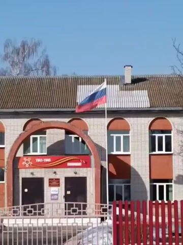 Here's a description of the video:

The video shows a two-story building with a Russian flag waving on a flagpole. The building appears to be made of brick with a brown shingled roof. Over the entrance to the building is a red and orange arch with the years "1941-1945" and the word "ПОМНИМ!" written on it. A fence is in the foreground. The sky is blue and clear.